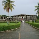 View of the unfinished and half-restored ancient temple in Hassan, Karnataka, showing weathered stone carvings and paused restoration work amidst green surroundings.