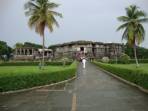 View of the unfinished and half-restored ancient temple in Hassan, Karnataka, showing weathered stone carvings and paused restoration work amidst green surroundings.